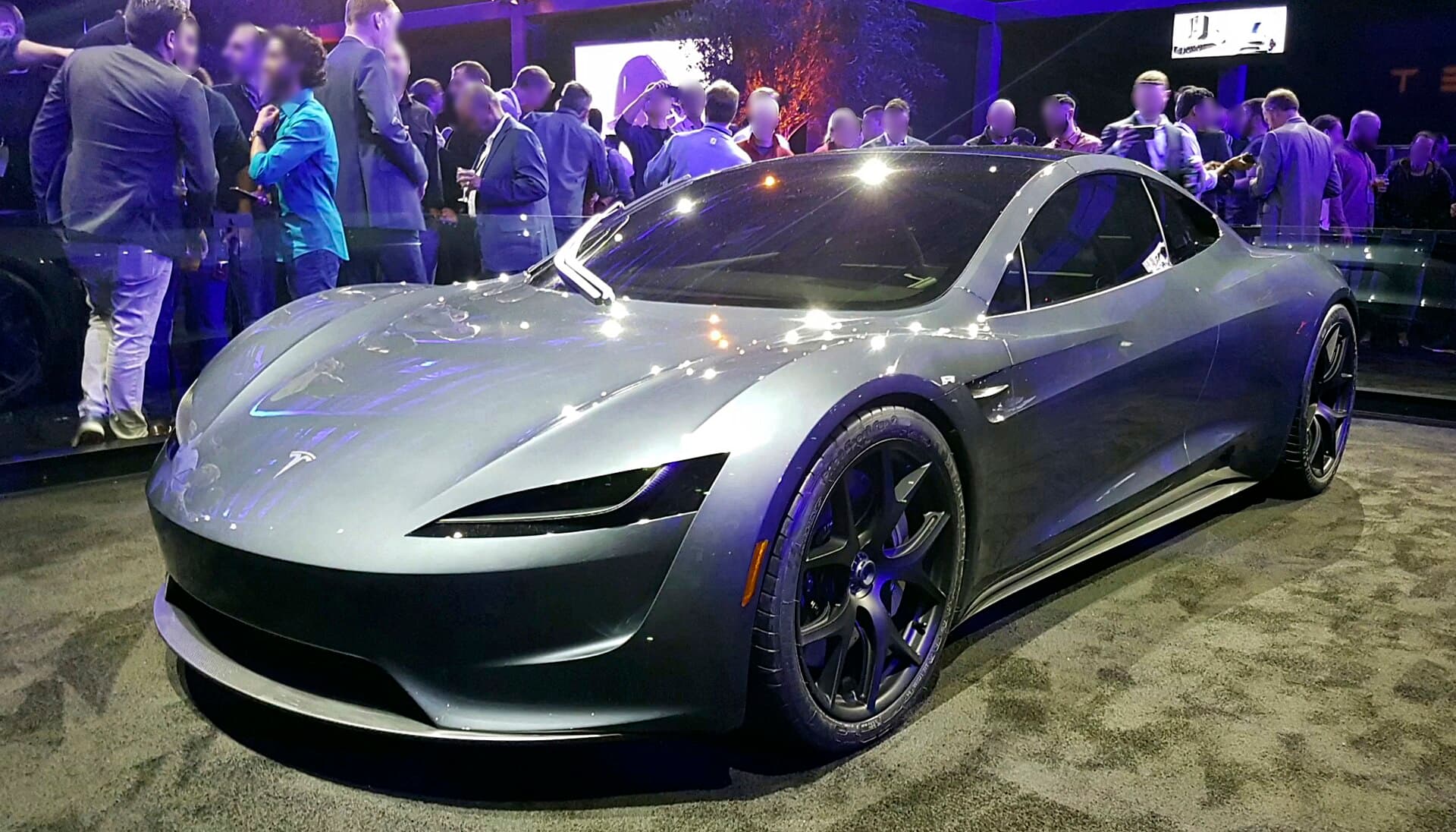 The Tesla Roadster prototype shown in 2017, silver, on display at an event with a crowd behind it; the new model looks different